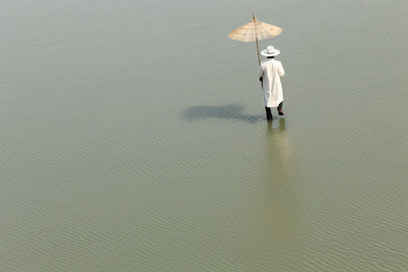 A solitary figure in a white coat, holding a traditional umbrella, walks amid shallow waters, creating a serene scene.の素材