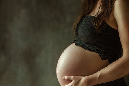 In a softly lit room, a pregnant woman cradles her rounded belly with one hand. The peaceful atmosphere highlights the beauty of motherhood and anticipation. Generative AI.の素材