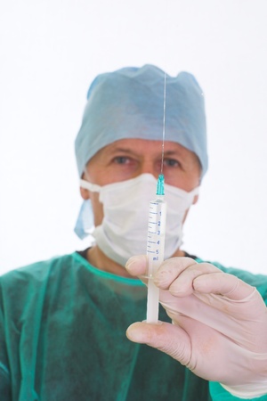 Image of a surgeon preparings an injection  He keeps the full syringe the needle up, removing a bubble of air  He wears rubber gloves, a mask, a cap and a green surgical coat の写真素材