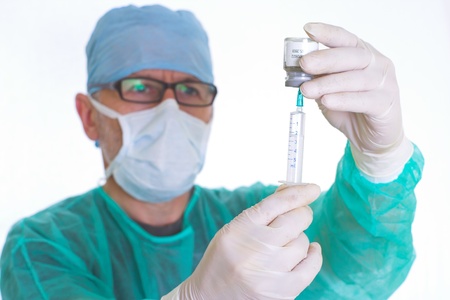 Image presents a doctor in green surgical smock, medical mask, medical bonnet and medical rubber gloves, who fills up the syringe, on white background の写真素材