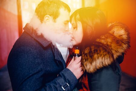 couple in love a man and a girl kiss and smoke electronic cigarettes vape mod and drip. There are clubs of steam. Concept smoking in public places.の写真素材