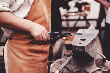 The smith is operating a hammer in a blacksmiths workshop on the anvil, holding the workpiece with pliers. Concept work on metal in the traditions.の写真素材