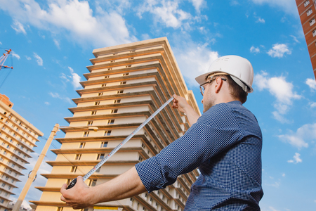 Male construction builder worker - foreman is an engineer in a protective helmet, glasses on the background of building a house with a crane Ruler measures. Concept calculation of construction time and cost.の写真素材
