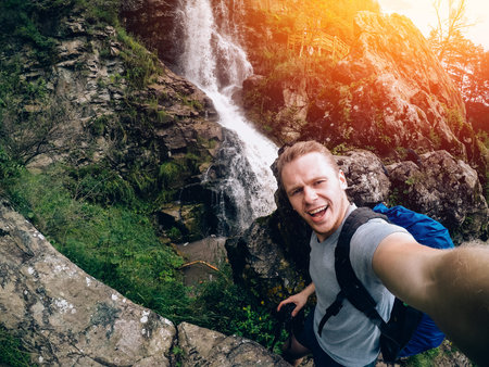 tourist man on a waterfall background holds an action camera and takes a picture of Selfie.
