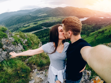 Guy and girl tourists are happy, hug and do selfie on action camera on background of mountains, forests. Concept kiss and travel. Russia, Altai Mountains.の写真素材