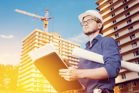 Close-up of a male construction worker wearing a helmet, glasses, a shirt in his hands holding a folder, papers, blueprints on the background of construction site: a crane, a multi-storey building.の写真素材
