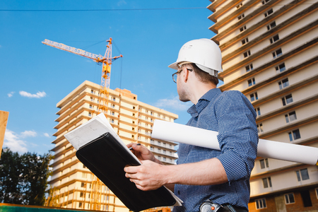 Close-up man builder European wearing helmet, glasses, shirt in his hands holding folder, papers, blueprints background construction site: crane, multi-storey house. Looks construction siteの写真素材