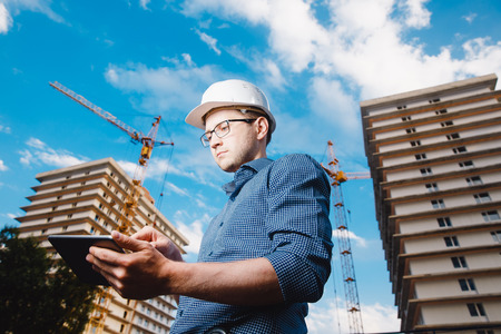 builder is a man in a protective helmet, glasses and a tablet computer in the hands of a crane, a multi-storey house and a blue sky. Concept construction, business, technologyの写真素材