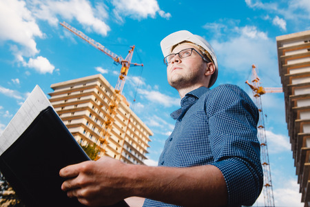 close-up man construction builder worker - foreman is engineer in protective helmet, glasses on the background of building house with crane. Concept calculation construction time, cost.の写真素材