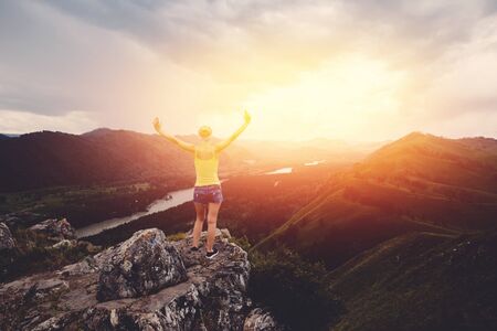 tourist girl in a plaid shirt and with a backpack standing on top of the mountain and having fun against the backdrop of mountains, rivers. The setting sun. Concept active rest. Space for textの写真素材