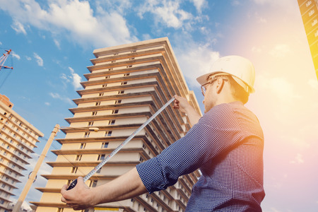 Male construction builder worker - foreman is an engineer in a protective helmet, glasses on the background of building a house with a crane Ruler measures. Concept calculation of construction time and cost.の写真素材