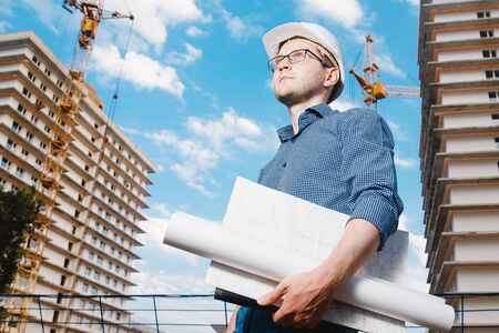 close-up man construction builder worker - foreman is engineer in protective helmet, glasses on the background of building house with crane. Concept calculation construction time, cost.の写真素材