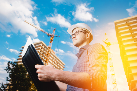 close-up man construction builder worker - foreman is engineer in protective helmet, glasses on the background of building house with crane. Concept calculation construction time, cost.の写真素材
