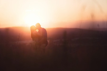 Lovers in mountains against background of fog and hills, a crimson, scarlet, red, orange sunset, sun shines in back, backlight, glare of sun. concept of first love and kiss of tender romanticの写真素材