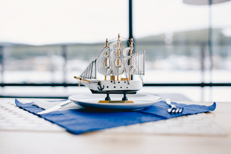 miniature sailing ship made of wood stands on a plate in a restaurant, behind it there is a window on the sea.の写真素材