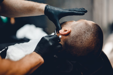 A man with a beard a client in a barbershop hairdresser on a shaving with a dangerous razor.の写真素材