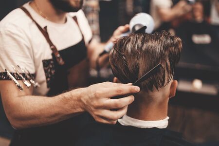 Close-up of a young hipster guy in a barbershop hairdresser cutting hair with scissors and a typewriter, waxing hair, blow-drying. Concept men's place, toned photo. Soft focus.の写真素材