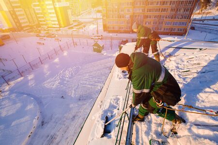 Snow cleaning. Team of male workers clean roof of building from snow with shovels in securing belts of mantra.の写真素材