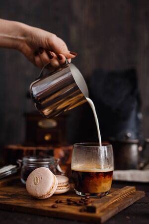 Coffee. Breakfast. Hand of girl pours milk into coffee, next to macaroni, in background coffee grinder, coffee beans, gray background, loft style. vertical frame.の写真素材