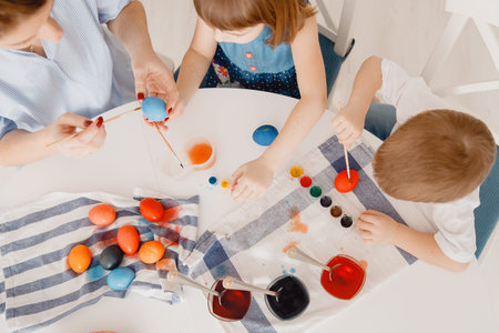 Mother, daughter and son paint eggs. White kitchen. A happy family is preparing for Easter. Top view