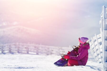 Snowboarding. girl is sitting hugging snowboard showing love of sport, in background of mountain in snow.の写真素材