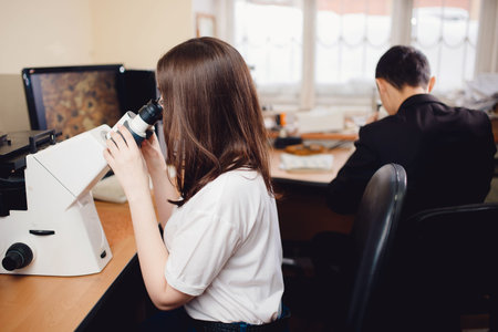 Student young woman sits behind a microscope in classroom, in background male student. Concept team work in scientific project.の写真素材