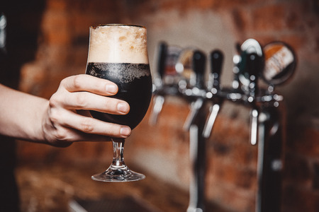 Barman holds in his hand dark unfiltered cold beer is poured into glass beaker against background of taps for bottling.の写真素材