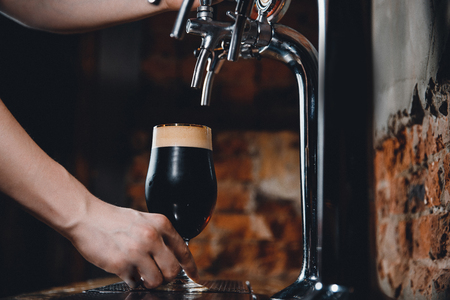 Dark unfiltered cold beer is poured into glass beaker, standing on background of taps for bottling.の写真素材
