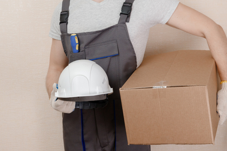 Man in uniform of delivery suit, loader, on his head white helmet, in hands box with loadの写真素材