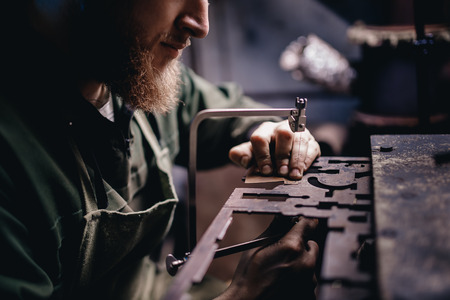 Man with beard in overalls saw with hand saw detail in garage workshop. Concept jewelery workの写真素材