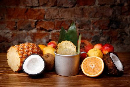 Refreshing tropical cocktail with pineapple, ice and alcohol in aluminum mug. In background, mix of exotic fruits is on display.の写真素材