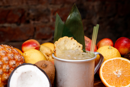 Refreshing tropical cocktail with pineapple, ice and alcohol in aluminum mug. In background, mix of exotic fruits is on display.の写真素材