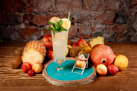 Refreshing cocktail with alcohol, mint, orange and ice in tall glass. Background, mix of fruits: pineapple, kiwi, lemon, pomegranate.の写真素材