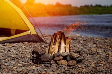 Bonfire is laid out of logs, it edged with stones, in background yellow tent on bank of lake, campingの写真素材