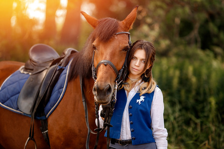 Young woman rider equestrian stands next to brown horse in forest.の写真素材