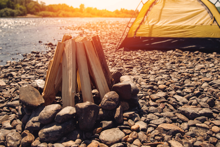 Bonfire is laid out of logs, it edged with stones, in background yellow tent on bank of lake, campingの写真素材