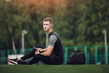 Young handsome man using phone while having exercise break in on football field with green lawn....