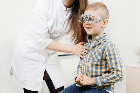 Small laughing boy with big blue eyes in special glasses for lens selection sits on chair office of ophthalmologistの写真素材