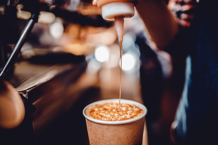 Author coffee, bartender before serving, pours caramel syrup, topping into finished drink in paper cup. Background is blurredの写真素材