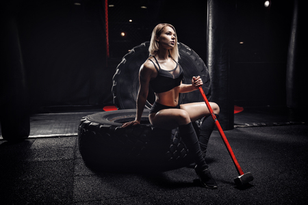 Beautiful athletic body of young woman holding hammer, sitting on tire wheel in gym. Workout fitness concept.の写真素材