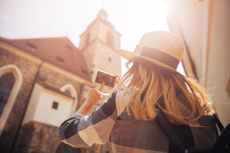 Young woman tourist in straw hat and with backpack is using phone as camera, taking pictures of buildings.の写真素材
