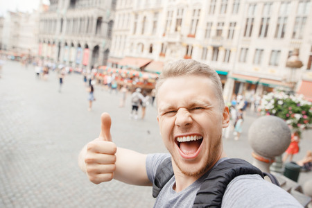 Young bearded blond guy smile traveler to Europe with backpack taking selfie on Grand place and showing Thumbs up thumb.の写真素材