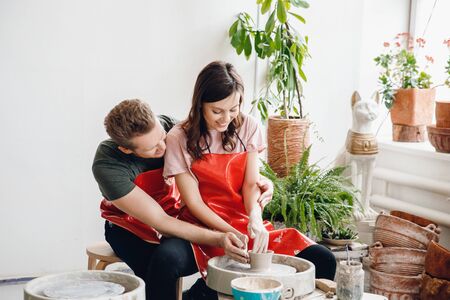 Tender embrace of romantic and young couple in red aprons behind potter wheel make ceramic dishes. Concept familyの写真素材