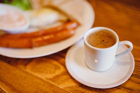 Classic English breakfast with espresso coffee in white mug, two sausages, fried eggs, toasts, lettuce on wooden backgroundの写真素材