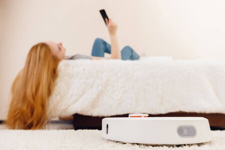 Young girl using automatic smart robot vacuum cleaner working on carpet. Controlling device with phoneの写真素材