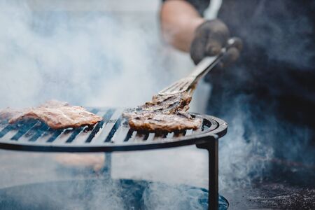 Cook grills steaks and pork ribs open fire. Turns meat over with tongs. Street food concept.の写真素材