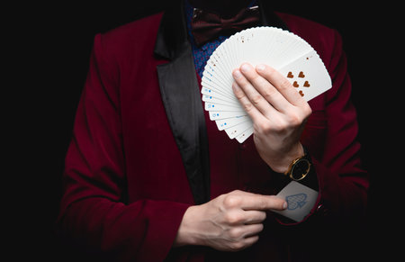 Illusionist man shows fan of playing cards takes ace out hand on black background.