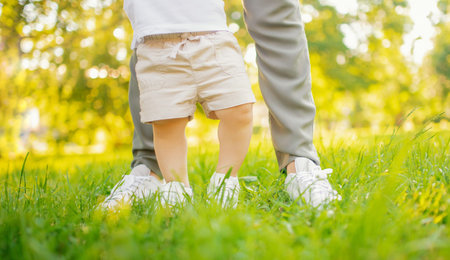Feet of child in white shoes take first step, mother of parent hedges and helps. Sunlight summer...