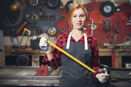 Beautiful young woman artisan engineer stands with ruler apron on background of craft tools. Concept small business in garage in industrial roomの写真素材