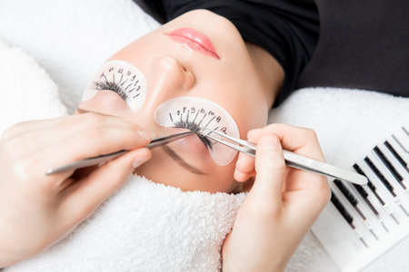 Eyelash extension procedure. Master tweezers sets fake lashes on beautiful woman